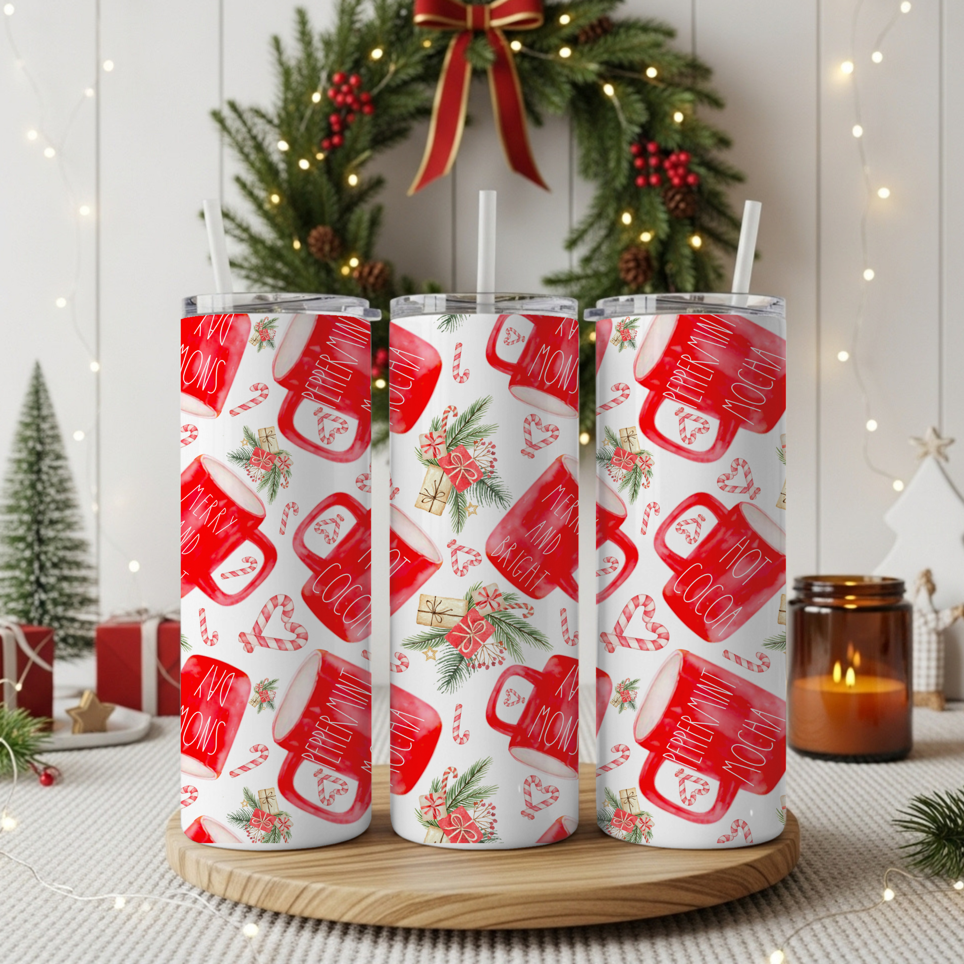 Three clear tumbler cups with white straws, printed with a red and white holiday mug pattern, displayed on a round wooden tray in a festive Christmas setting with a wreath, candles, and lights.
