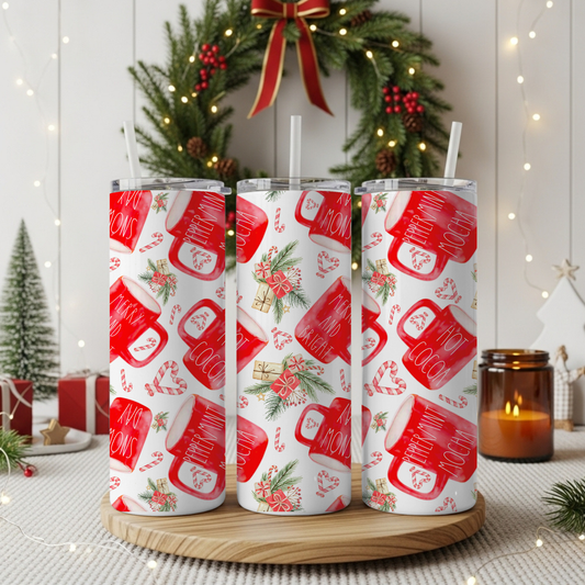 Three clear tumbler cups with white straws, printed with a red and white holiday mug pattern, displayed on a round wooden tray in a festive Christmas setting with a wreath, candles, and lights.