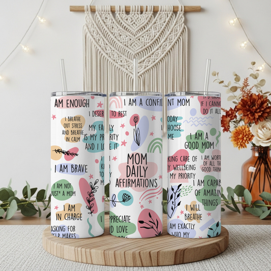 Three white insulated tumblers with metal lids and clear straws, standing on a round wooden tray. The tumblers feature colorful affirmation quotes and boho-style illustrations in pink, teal, mustard, and brown.