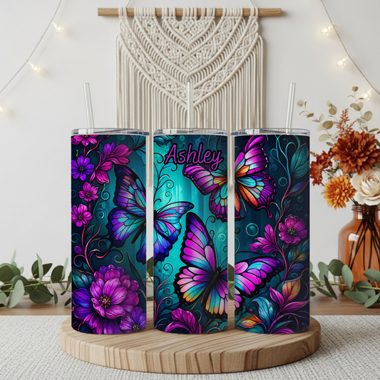 Three slim stainless steel tumblers with clear straws, decorated with vibrant purple, pink, and blue butterfly and floral designs on a dark teal background. The center tumbler features the name 'Ashley' in pink script.