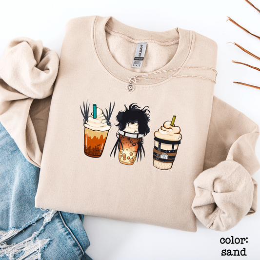 Flat lay of a sand-colored graphic sweatshirt featuring three spooky Halloween-themed iced coffee cups side-by-side with funny character details.