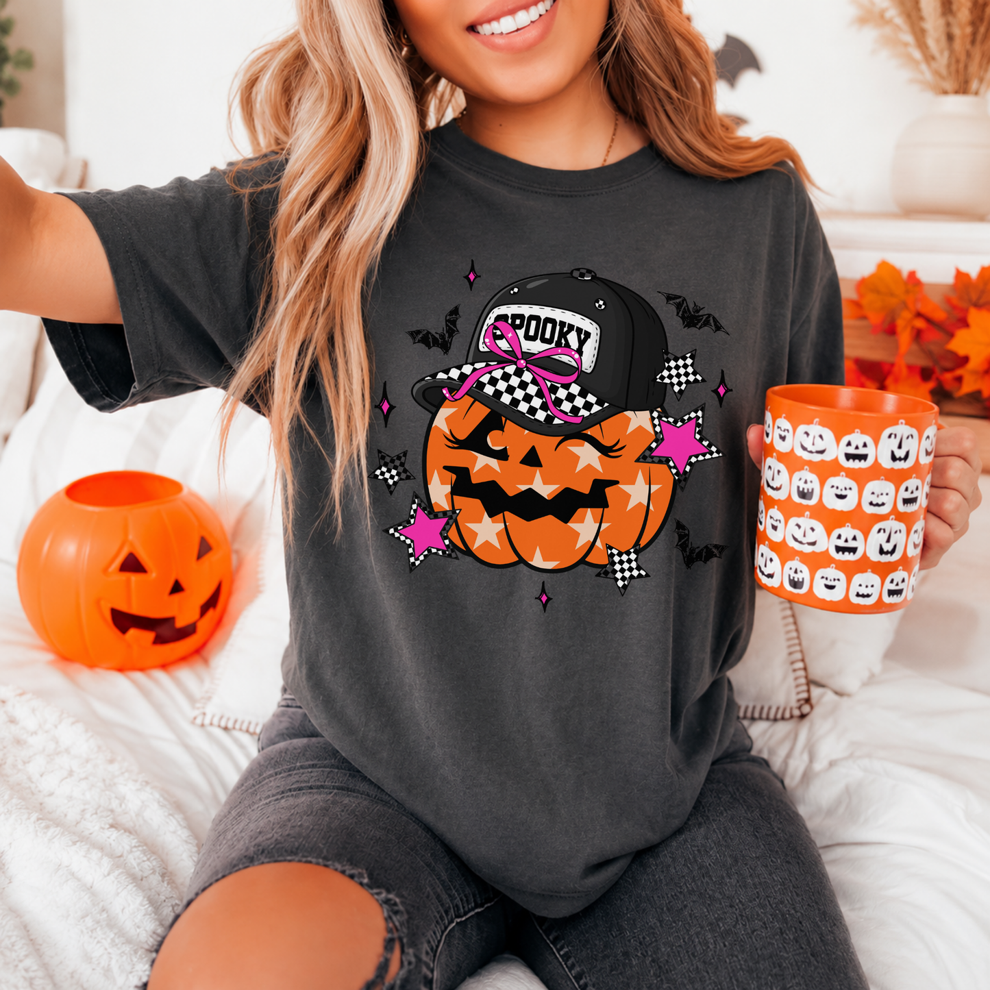 Person wearing a black t-shirt with Halloween-themed design, holding an orange mug with pumpkins, in a cozy indoor setting.